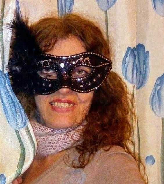 Maschera nera con dettagli scintillanti e piume, donna con capelli ricci rossi sorridente, decorazioni festose, stile carnevale o party, sfondo con disegni di fiori blu.