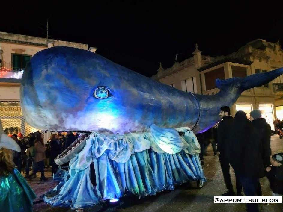 Gigantesca balena decorativa di carta durante una festa in strada, illuminata di blu, con persone che la osservano e la fotografano, atmosfera festiva e ricca di colori notturni, evento pubblico, carri di carnevale, decorazioni di strada.