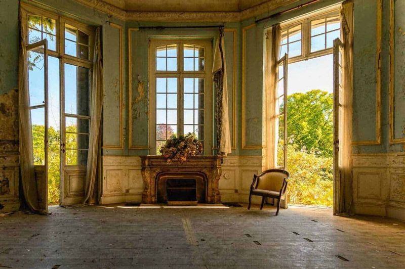 Città medievale XI secolo tra mura fortificate: aspetto e vita quotidiana 8 Ancora di una stanza di lusso abbandonata, con grandi finestre che lasciano entrare luce naturale, pareti decorate e un camino antico con fiori, in un ambiente ricco di storia e decadimento.