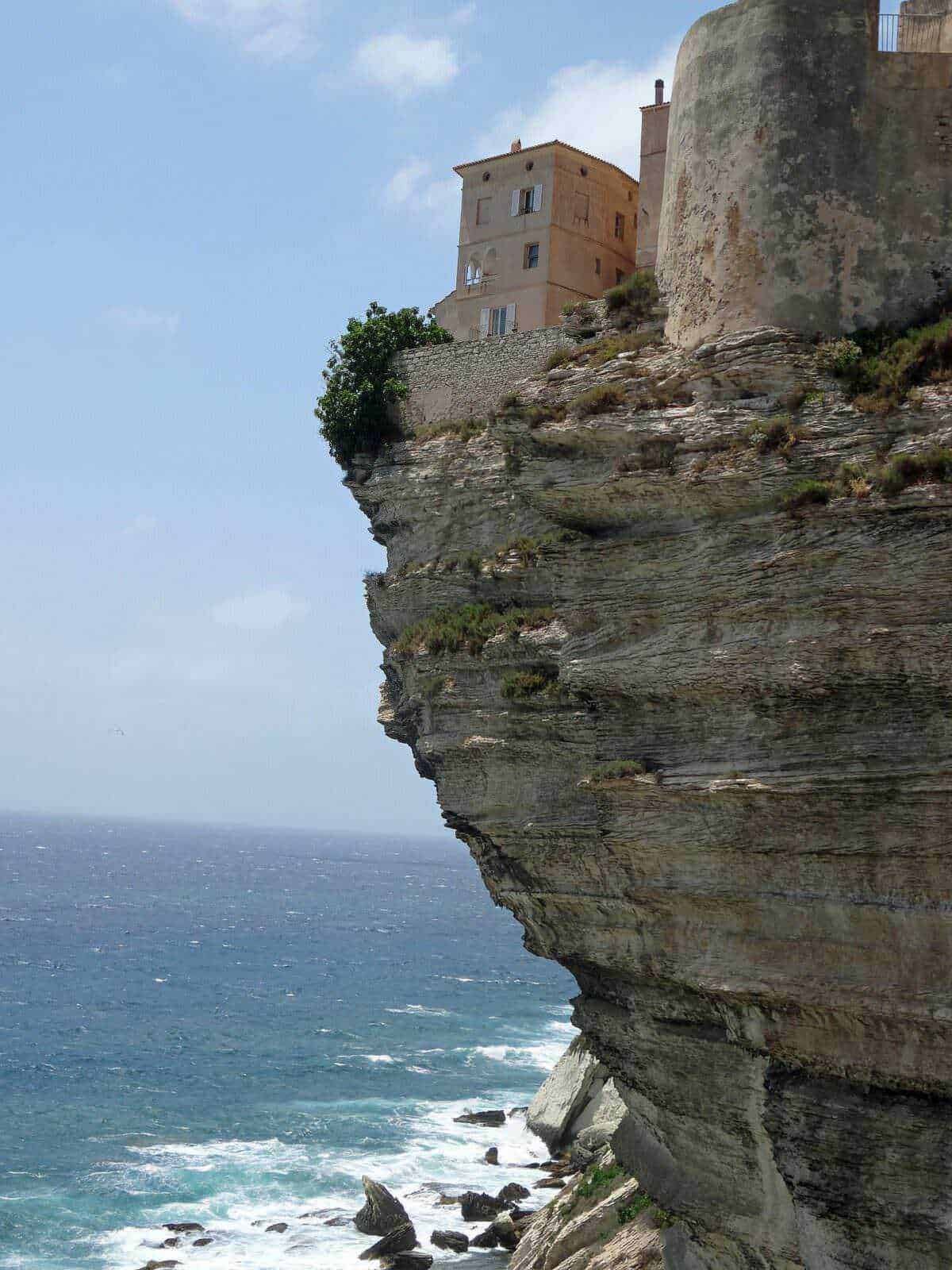 Cosa vedere in Corsica. Ingresso di un antico edificio in cima a una scogliera che si affaccia sul mare, con pareti rocciose a picco e onde che si infrangono sulla riva sottostante. Un paesaggio mozzafiato sulla costa italiana.