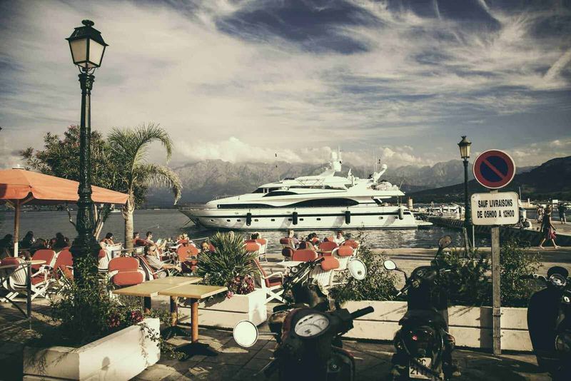 Come arrivare in Corsica da Roma, info viaggi (2026) 2 Faro di pietra lungo il porto con barche di lusso e motoscooter, atmosfera rilassante con persone sedute ai tavoli, montagne sullo sfondo, cielo nuvoloso, segnale di divieto di sosta.