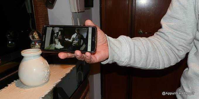 Ripresa di una mano che tiene uno smartphone con schermo acceso, inquadrando una scena di una conversazione o video alla luce di una lampada d'ambiente.