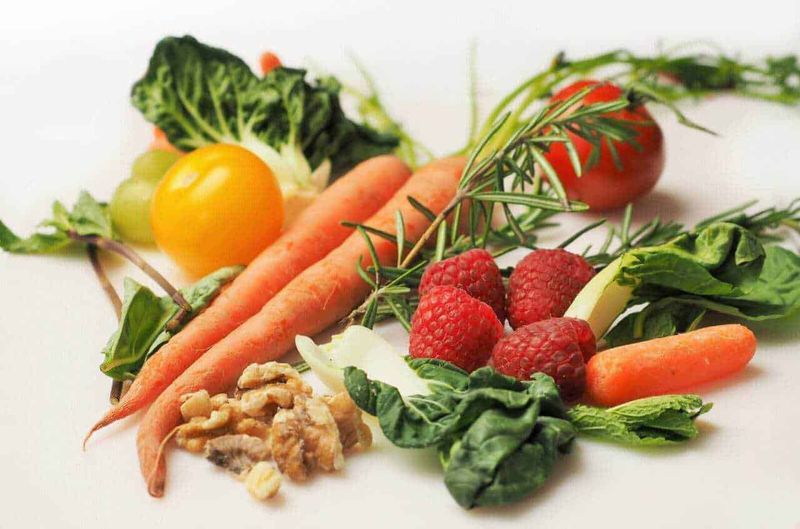 Pomodori, carote, lamponi, insalata e noci, alimenti freschi e salutari disposti su sfondo bianco, ideali per ricette di cucina naturale e sana.