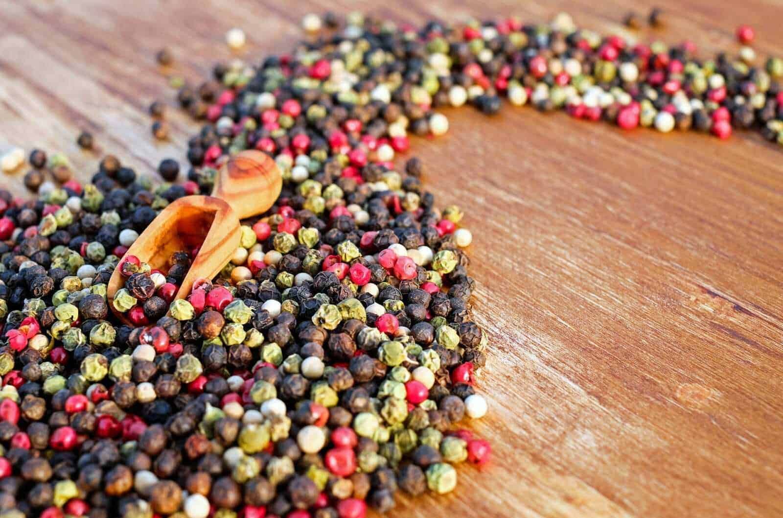 Peppercorns misti di vari colori sparsi su un tavolo di legno con cucchiaio in legno, condimento naturale per piatti e cucina internazionale, focus su spezie ed erbe aromatiche.