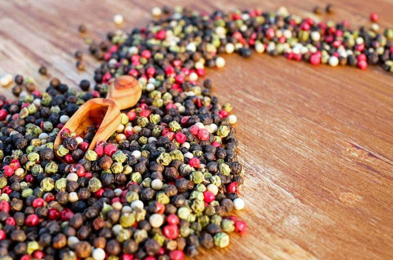 15 rimedi naturali per controllare l'ipertensione 4 Peppercorns misti di vari colori sparsi su un tavolo di legno con cucchiaio in legno, condimento naturale per piatti e cucina internazionale, focus su spezie ed erbe aromatiche.