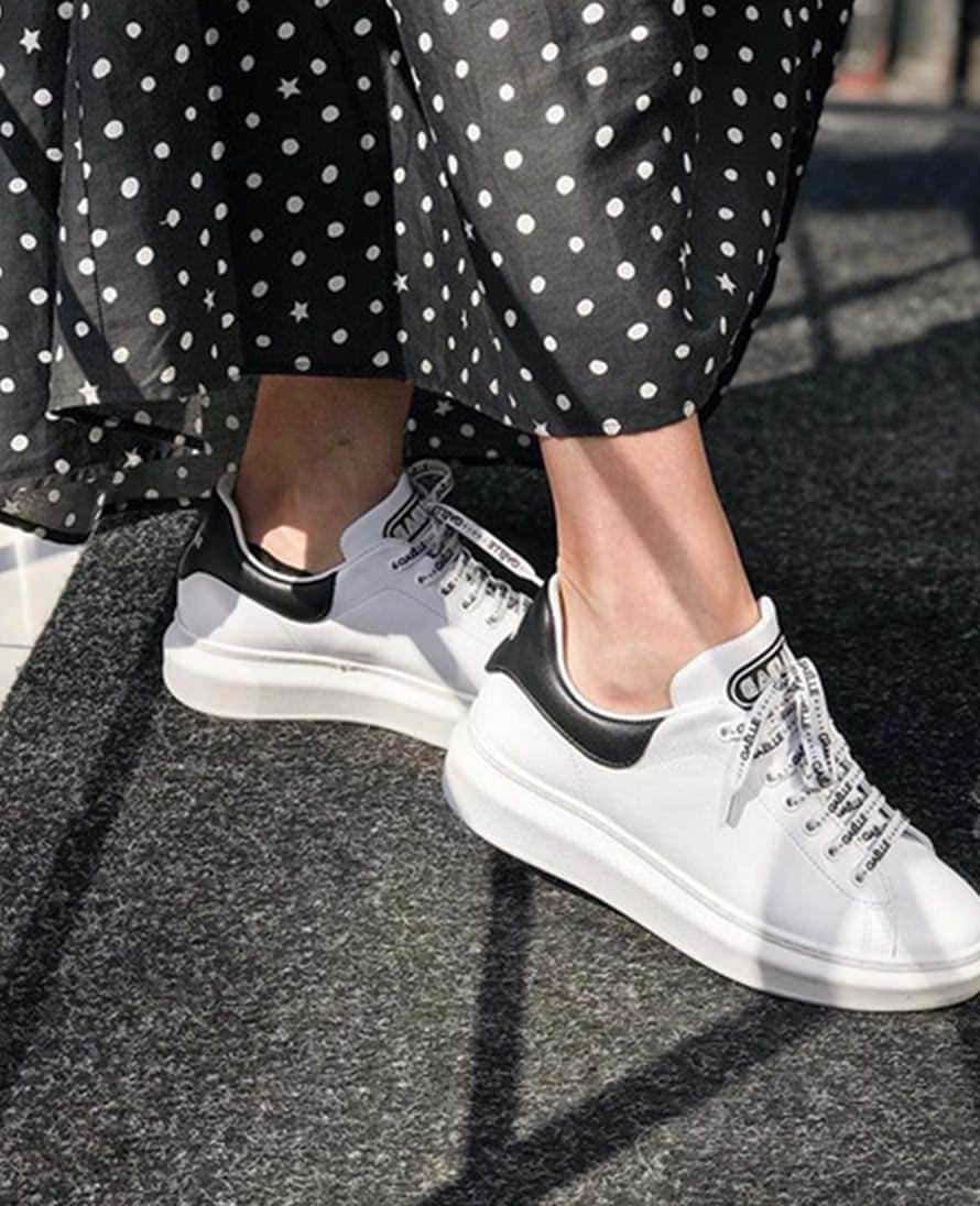 Scarpe da sneaker bianche con dettaglio nero, indossate con pantaloni a pois neri e bianchi, appoggiate su un pavimento in pietra, stile casual e alla moda.