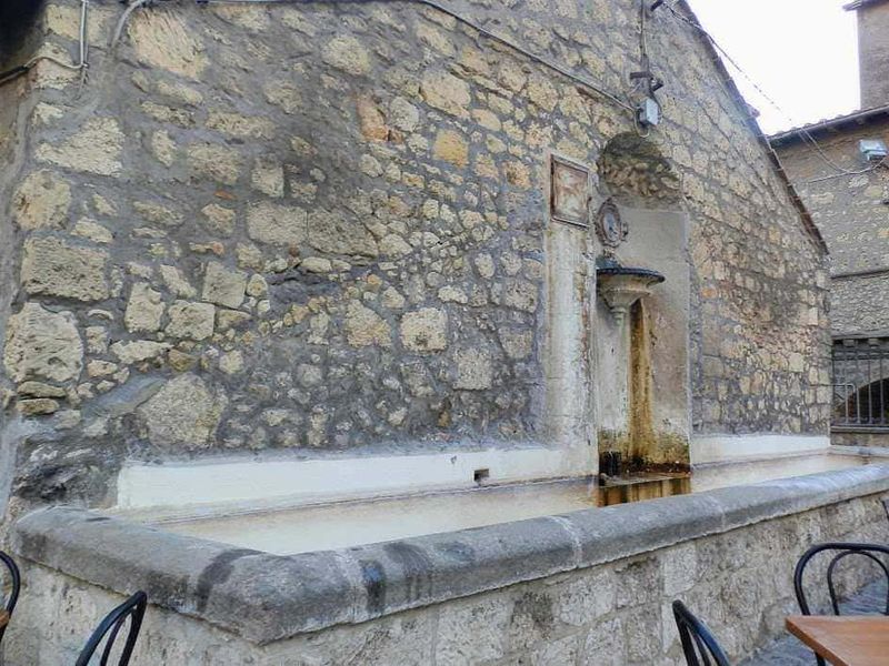 Vecchio fontanile in pietra con abbeveratoio in marmo, situato in un borgo storico italiano, caratterizzato da pareti in pietra e archi tradizionali, ideale per fotografie di paesaggi rurali e architettura storica.