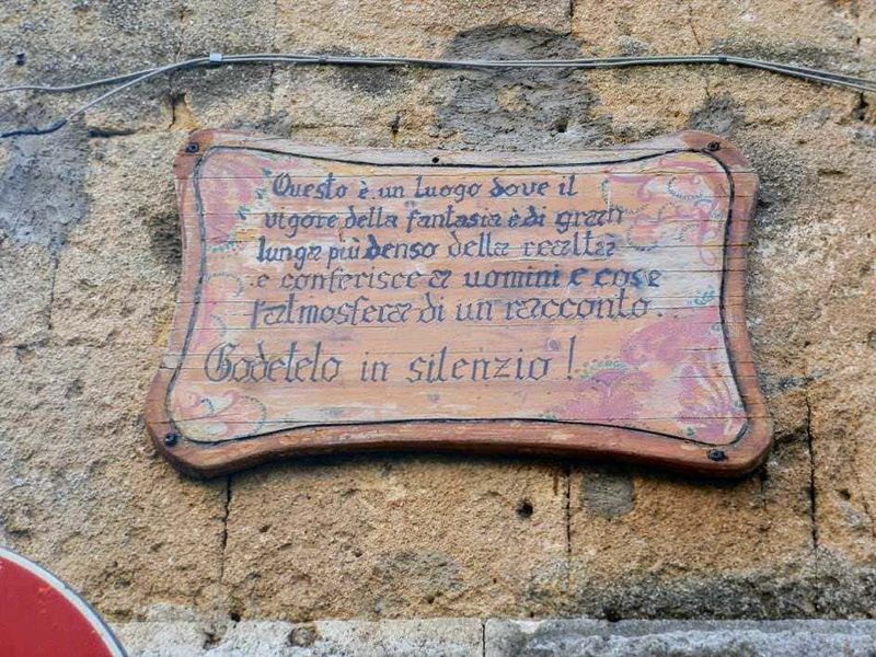 Un cartello in legno appeso a una parete in pietra con citazione poetica sulla fantasia e la realtà, in stile rustico e artistico.