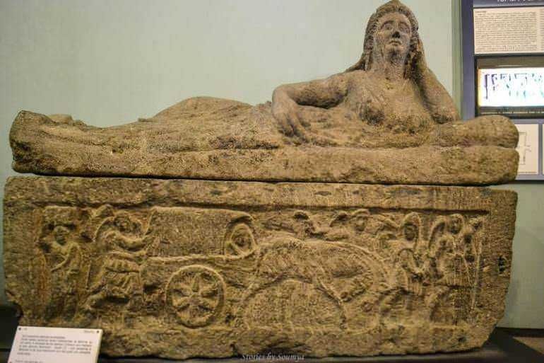 Antico sarcofago in pietra con rilievo di carri e figure mitologiche, risalente all'epoca romana, esposto in museo storico. Perfetto esempio di arte funeraria romana e simbologia religiosa.