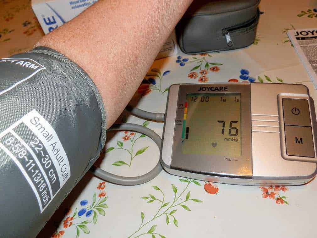 Pulsossimetro digitale che misura la saturazione di ossigeno nel sangue, visualizzata come 76%, collegato a un braccio e posizionato su un tavolo con decorazioni floreali, durante una misurazione a casa.