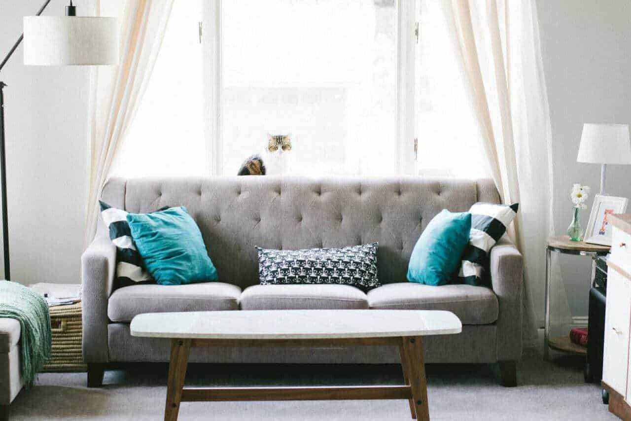 Atmosfera accogliente in soggiorno con divano tondo, cuscini blu e decorazioni moderne su tavolino e scaffale, grande finestra con tende leggere, perfetta per ambienti di stile e comfort.