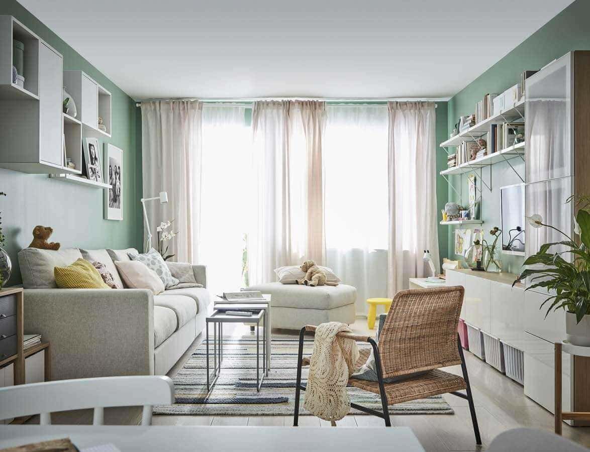 Spazio soggiorno luminoso con pareti verdi, divano beige, librerie con libri e decorazioni, grande finestra con tende bianche, arredamento accogliente e moderno.