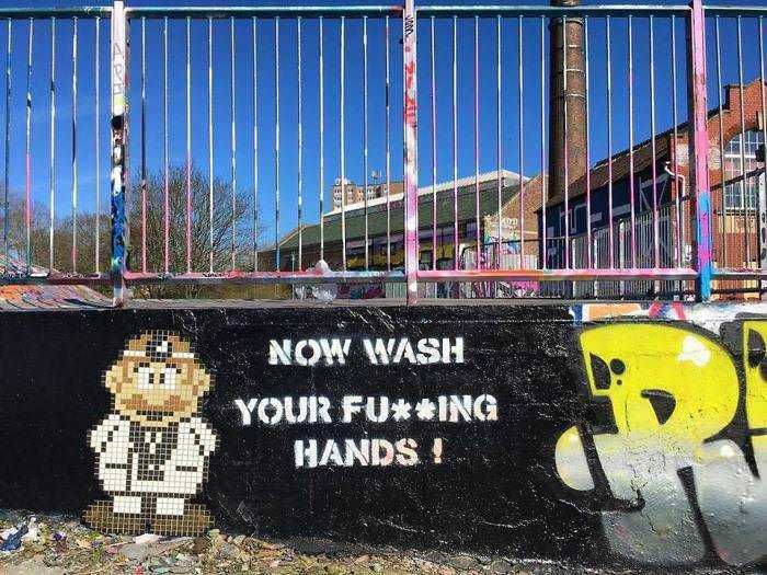 Lavagna con graffiti e scritta "Now wash your f***ing hands" in un'area pubblica, attività di street art e street culture in contesto urbano.