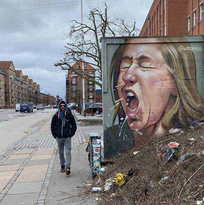 Vandalo street art con murale di una donna che urla, sulla strada di una città europea, con un uomo vestito casual, cielo grigio e edifici in sfondo.