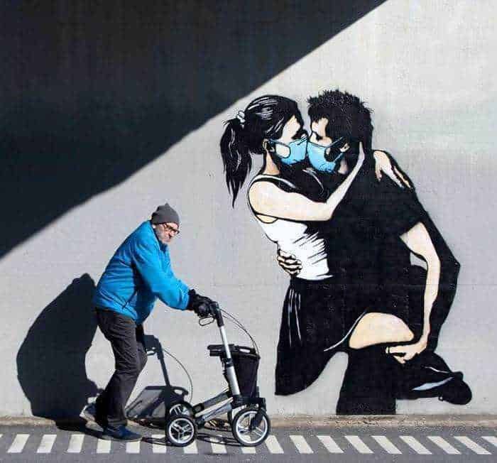 Ragazzo anziano con deambulatore che passa davanti a una grafica di due persone che si abbracciano indossando mascherine, street art in ambiente urbano, riflesso del periodo pandemico, simbolo di affetto e distanza sociale.
