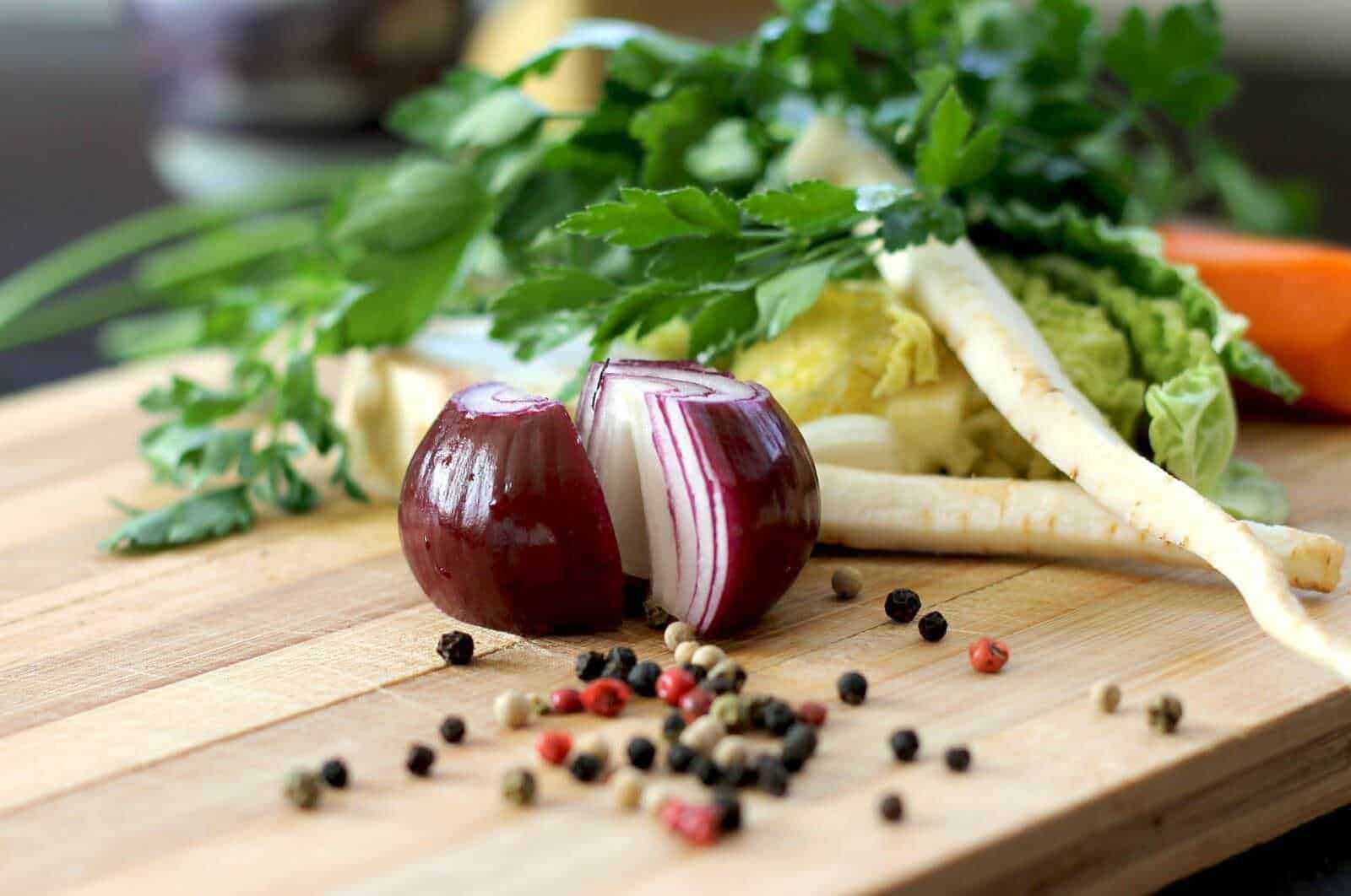 Verdure fresche e spezie su tagliere di legno, con cipolla rossa, prezzemolo, foglie di insalata e pepe nero, ideali per ricette leggere e salutari.