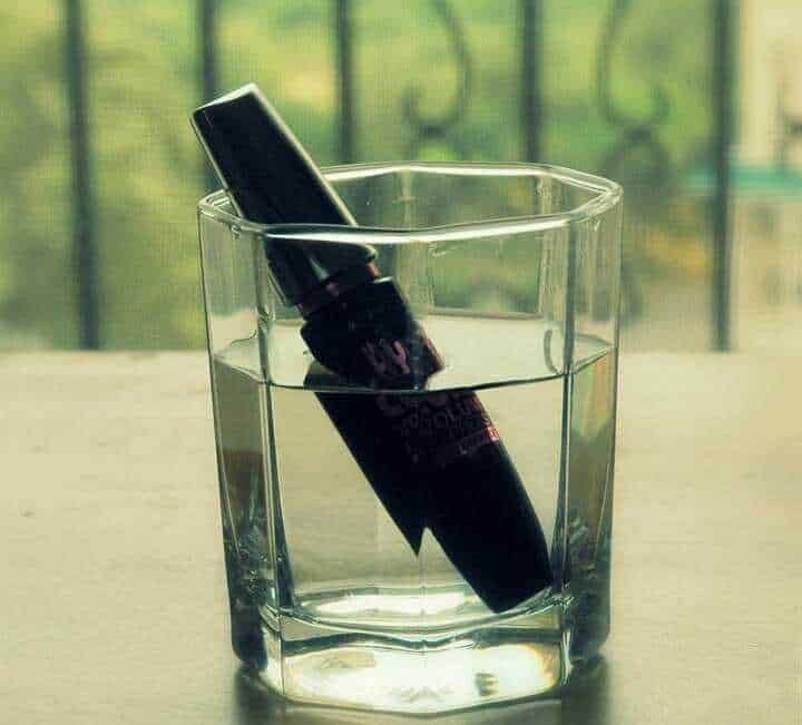 Penna a sfera inchiostro nero immersa nell'acqua in un bicchiere trasparente, simbolo di scrittura e creatività, con sfondo naturale.