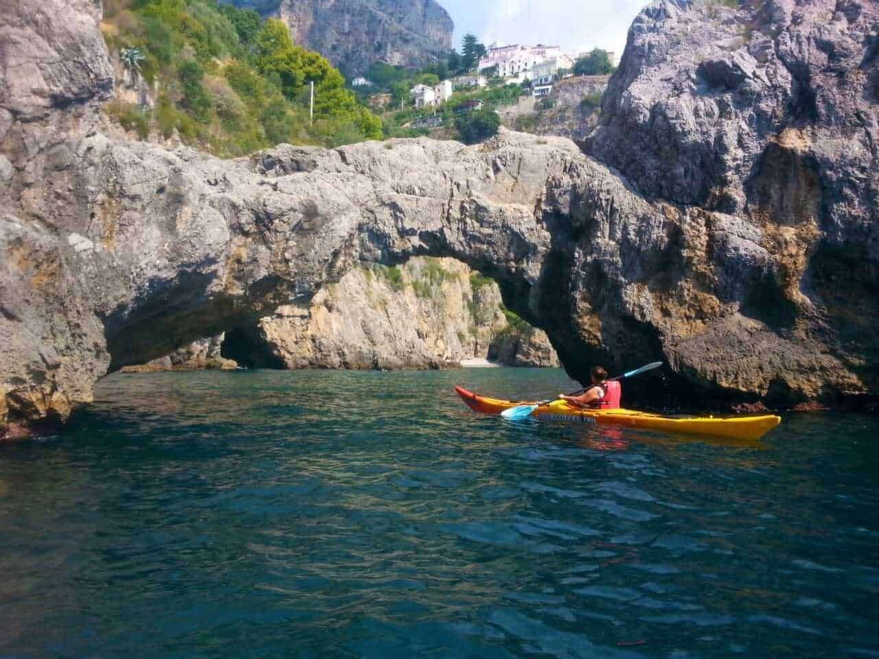 Kayak sotto un arco roccioso naturale nel mare con paesaggi montani e case in background, ambientazione estiva, attività all'aperto, avventure acquatiche, turismo naturale, escursioni in kayak.