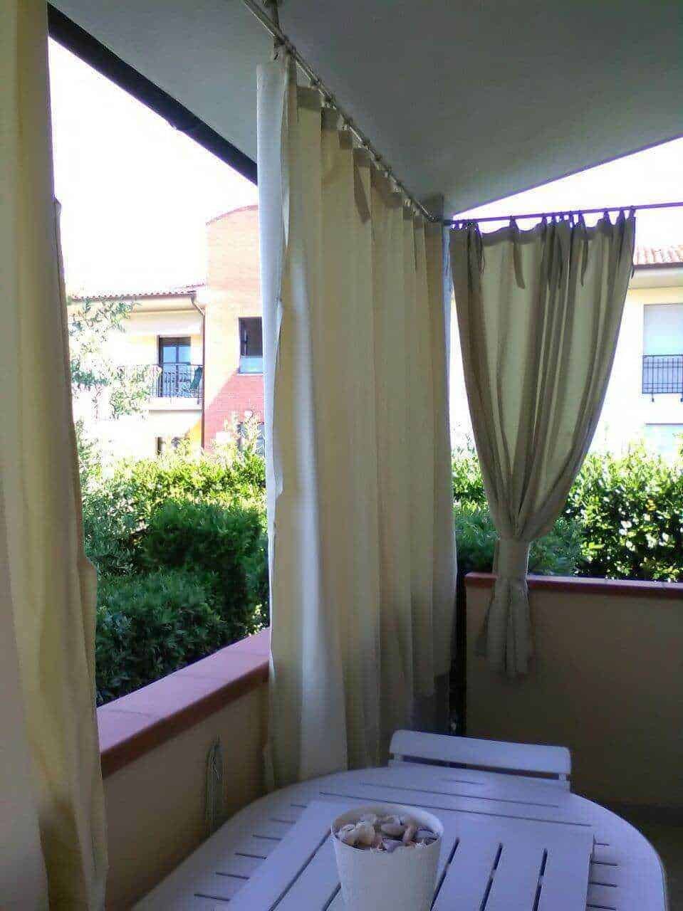 Balcone di un appartamento con tende chiare e vista su giardino e edifici residenziali, ambiente tranquillo e soleggiato, ideale per momenti di relax all'aperto.