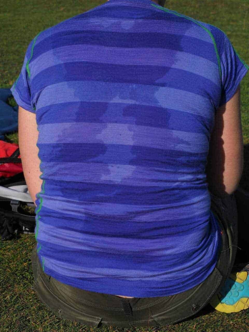 Maglietta sportiva azzurra a righe, vestita da una persona seduta all'aperto su un prato verde con attrezzature sportive sullo sfondo.