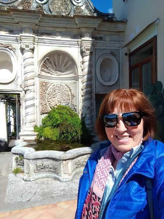 Fonte: Appunti Sul Blog, immagine di una donna sorridente con occhiali da sole in un cortile storico con fontana e decorazioni in stile barocco.