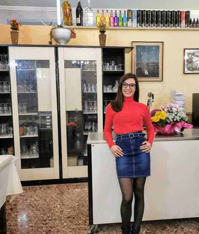 Vetrina di bar con bicchieri e bottiglie sullo sfondo, donna sorridente in abito rosso e gonna in denim, decorazioni floreali e arredo interno, immagine ideale per promozione bar e ambienti conviviali.