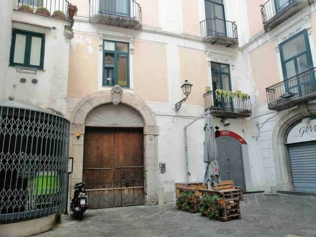 Rustic urban street scene with old building facades, balconies with potted plants, vintage street lamp, wooden door, scooter, and outdoor seating area with pallet furniture in Italy.