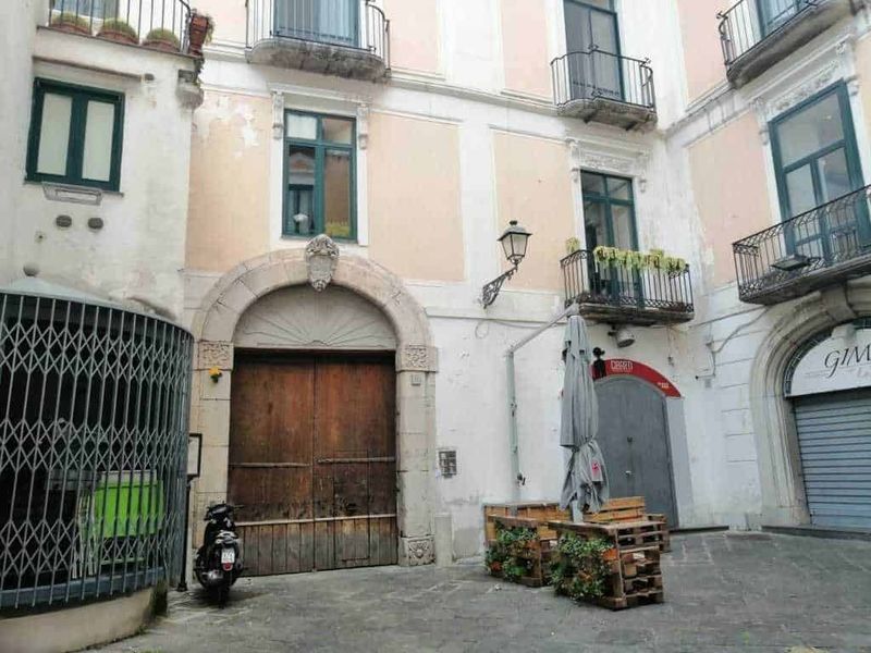 12 Edifici longobardi a Salerno (2026), da Palazzo d'Avossa: stile barocco, coppedè e storia medievale 11 Rustic urban street scene with old building facades, balconies with potted plants, vintage street lamp, wooden door, scooter, and outdoor seating area with pallet furniture in Italy.