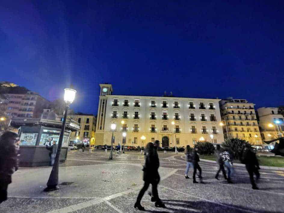 Pontile illuminato in piazza a notte, edifici storici, persone che camminano, atmosfera urbana, piazza vivace, luci della città, scena notturna, architettura storica, vita cittadina, movimento e socialità, Appunti Sul Blog.