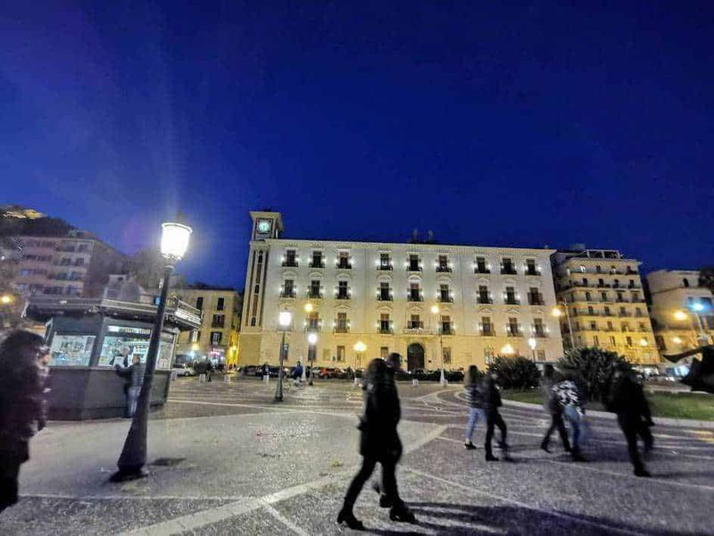12 Edifici longobardi a Salerno (2026), da Palazzo d'Avossa: stile barocco, coppedè e storia medievale 4 Pontile illuminato in piazza a notte, edifici storici, persone che camminano, atmosfera urbana, piazza vivace, luci della città, scena notturna, architettura storica, vita cittadina, movimento e socialità, Appunti Sul Blog.