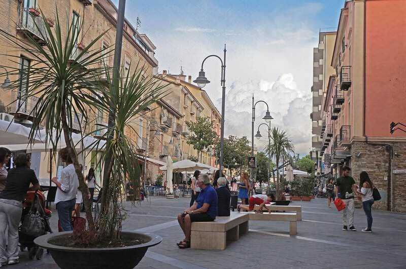 Vita urbana sul lungomare con panchine, palme e persone che passeggiano, atmosfera rilassante, negozi e caffè, ideale per immagini di lifestyle, turismo e divertimento in città.