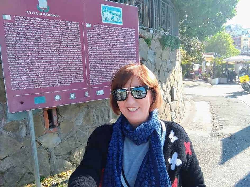 Vivace donna con occhiali da sole e sciarpa blu, sorridente davanti a un tabellone informativo su Agropoli, in una zona all'aperto al sole con alberi e strutture nel background.