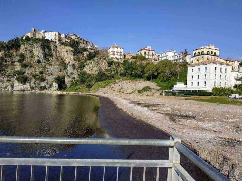 Ferrovia e case storiche sulla scogliera costiera di Sorrento, vista dall'alto di una passeggiata sulla spiaggia, con mare calmo e cielo sereno, in un tipico paesaggio italiano.