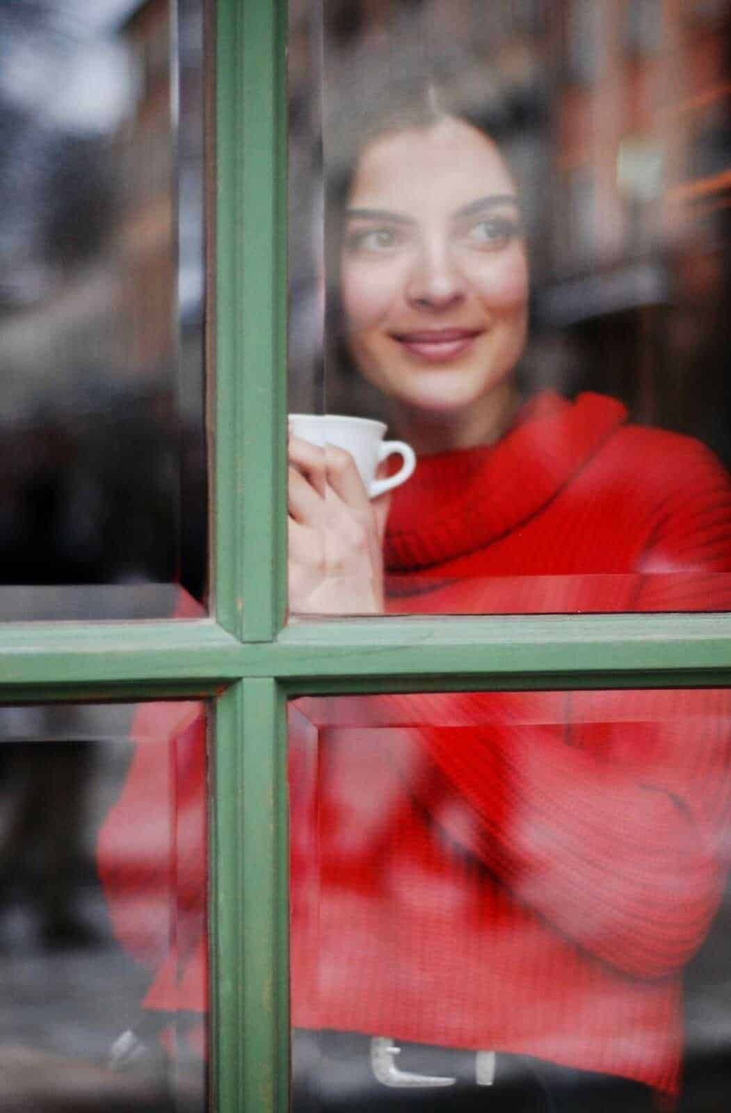 Tiziana sorridente guarda fuori da una finestra mentre sorseggia una tazza di caffè, creando un momento di relax e intimità in un ambiente accogliente.