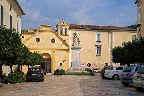 Piazza storica con monumento e chiesa antica in un centro urbano italiano, parcheggio e alberi, atmosfera tranquilla e suggestiva, ideale per turismo e cultura, in un'incantevole località italiana.