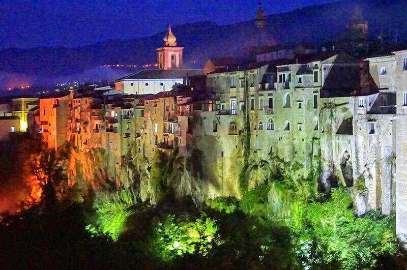 Sant'Agata de' Goti chiese ed eventi della Perla del Sannio 17 Palazzi colorati e case storiche su una scarpata, illuminati con luci verdi e arancioni, con sfondo di colline e cielo notturno, rappresentano un affascinante paesaggio urbano italiano.
