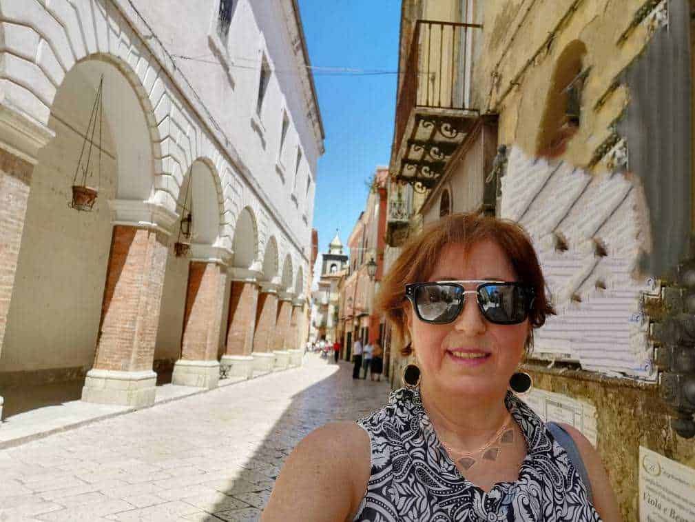 Scorcio di strada storica con arconi in mattoni e donna con occhiali da sole che prende un selfie a lume di sole.