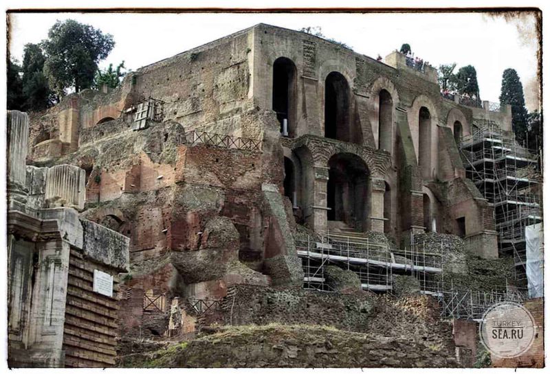 Come vivevano i Romani la vita quotidiana (2026) 3 Antica rovina archeologica con resti di un edificio storico, probabilmente parte di un sito romano o antico, con scale di accesso e scavi archeologici, circondata da vegetazione e scaffalature di restauro.