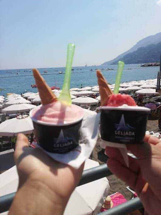 Gelato artigianale in coppa con cono, godendo di una vista mare con ombrelloni e montagne sullo sfondo, ideale per momenti di relax e vacanza al mare.