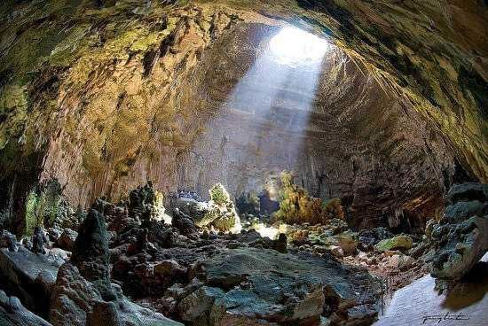 Caverna naturale con luce solare che entra dal soffitto, rocce e terra nel interno di una grotta, paesaggio sotterraneo suggestivo e misterioso.