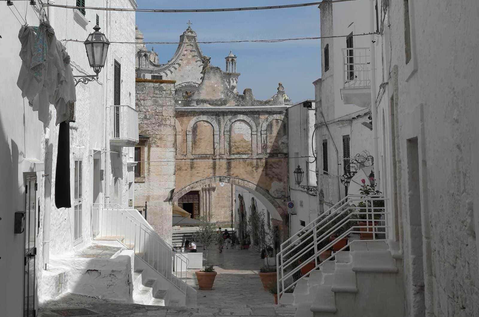 Vecchia strada di un paese in Puglia con mura bianche e archi, chiesa storica sullo sfondo, atmosfera mediterranea e architettura tradizionale.