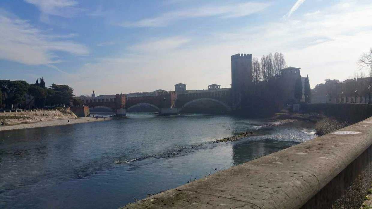 Ponte di Castelvecchio sul fiume Adige a Verona, storico ponte medievale con torre e archi, panorama urbano, zona pedonale lungo il fiume, cielo sereno e riflessi sull'acqua, punti di interesse turistico.