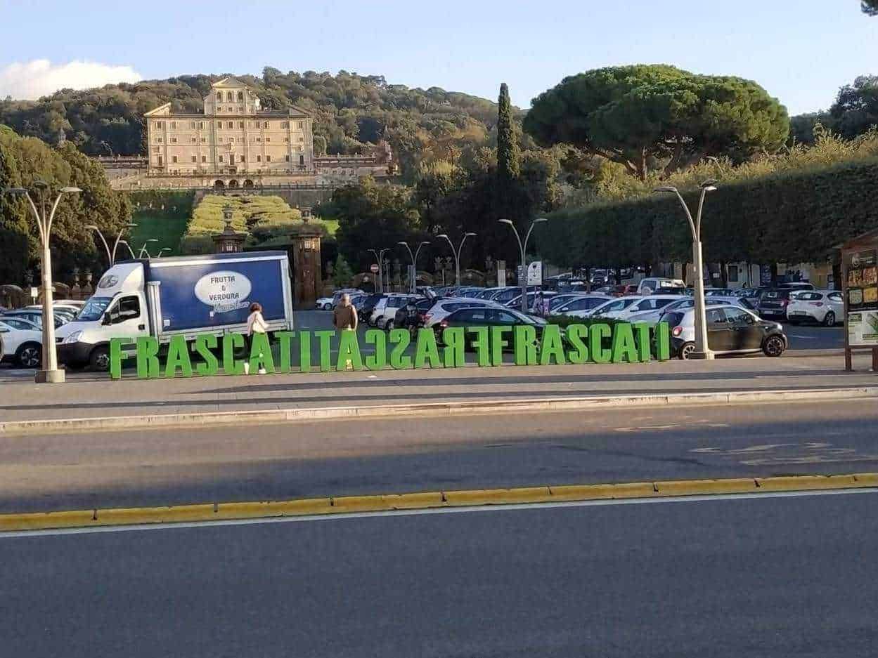 FRASCATI in lettere grandi verdi davanti a un parcheggio con auto e un camion, vista sul paesaggio con colline e un grande edificio storico sullo sfondo, zona urbana e naturale, Italia.