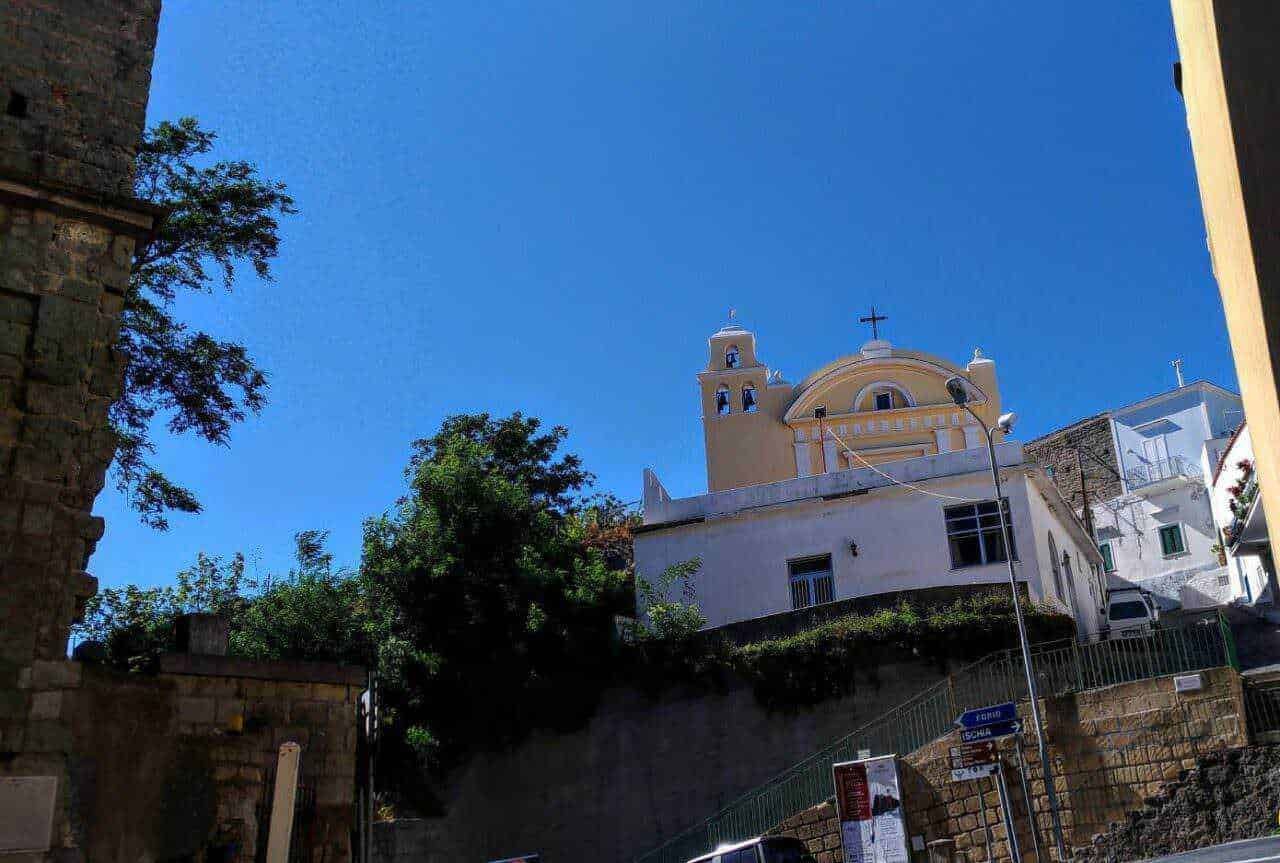 Villa storica su collina con chiesa barocca sotto un cielo azzurro, contorni di case bianche e verde foliage, atmosfera mediterranea, in un paesaggio urbano di Napoli, Italia.