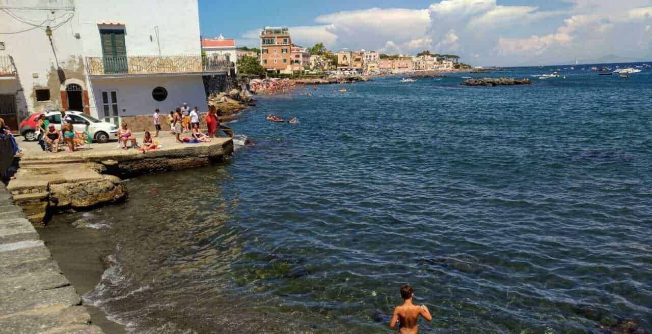 Spiaggia affacciata sul mare con persone che gustano il panorama, ombrelloni e costruzioni colorate nello sfondo, atmosfera estiva e rilassante in un borgo marino italiano.