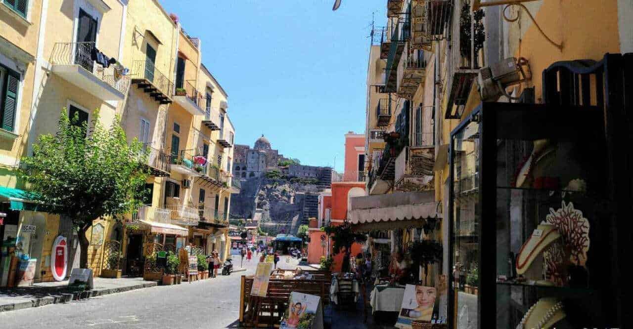 Colorata via di Napoli con edifici storici, negozi e ristorante all'aperto sotto un cielo azzurro, con il Castel dell'Ovo sullo sfondo, rappresenta il fascino di una città mediterranea.