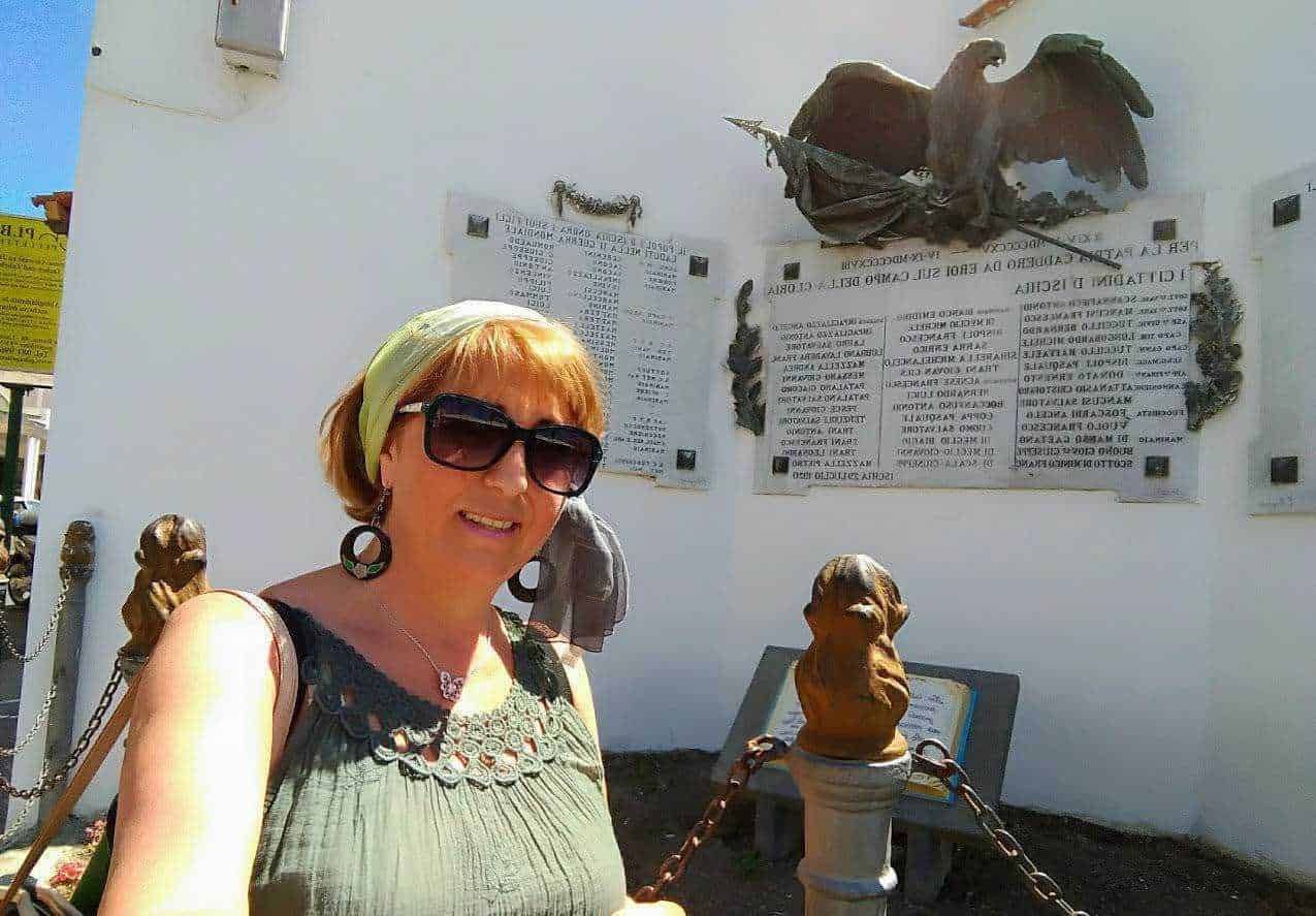 Femminile turista con occhiali scuri e foulard seduta davanti a monumento commemorativo di guerra in Italia.