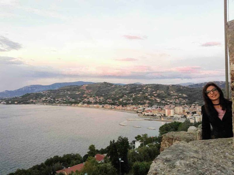 Il paesaggio del mare, come si chiama la costa 2 Panorama del mare e della costa di Napoli al tramonto, con una donna sorridente seduta su un muro di pietra, sullo sfondo le colline e il cielo colorato.