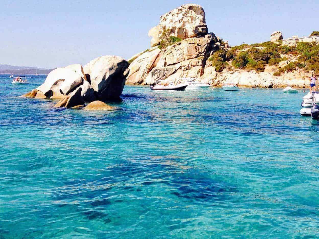 Spiaggia con acque cristalline e scogliere di granito sullo sfondo, ideale per fotografie di viaggio e vacanze al mare, con barche e natura in primo piano.