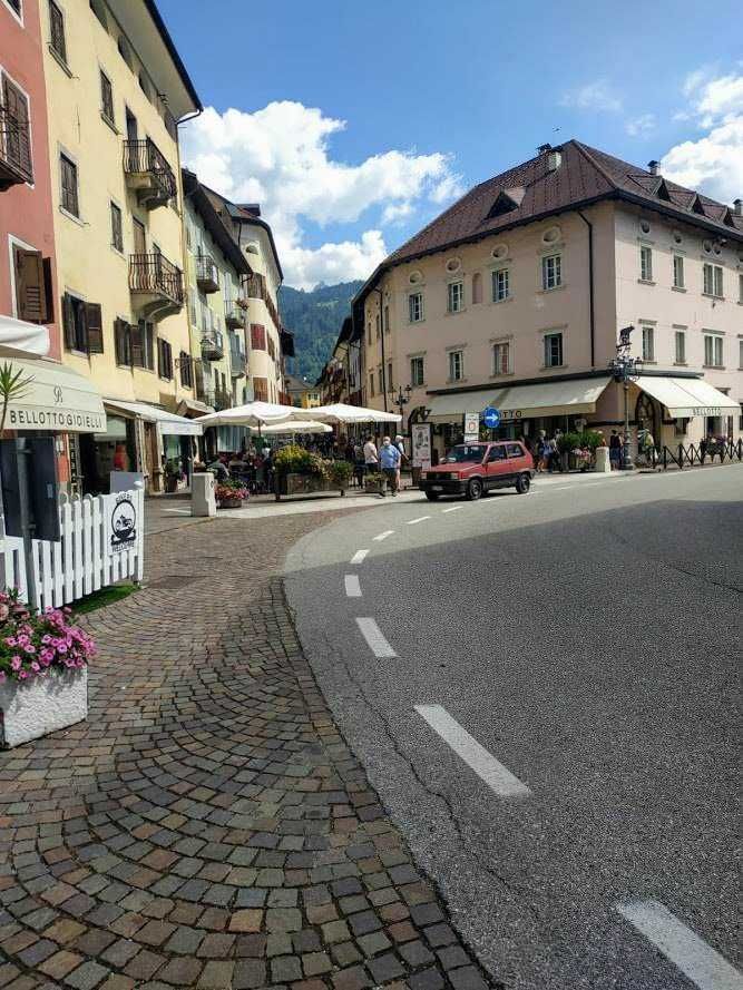 Stretta strada di montagna con edifici colorati e caffè all'aperto, turisti che passeggiano e auto d'epoca, atmosfera affascinante di un borgo alpino in giornata soleggiata, con montagne sullo sfondo.